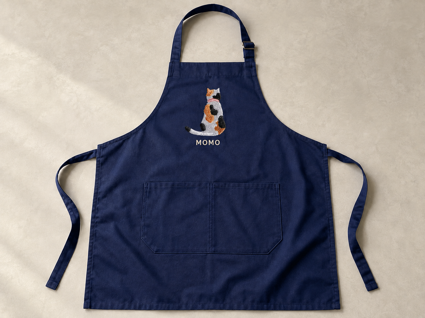 ネイビーのエプロンの胸元に、三毛猫の後ろ姿と MOMO の名前を刺繍したもの